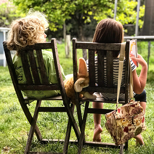 Mentalcoaching Einzeltraining: Rückansicht von zwei Kindern, die in einem Garten auf zwei Gartenstühlen aus Holz sitzen